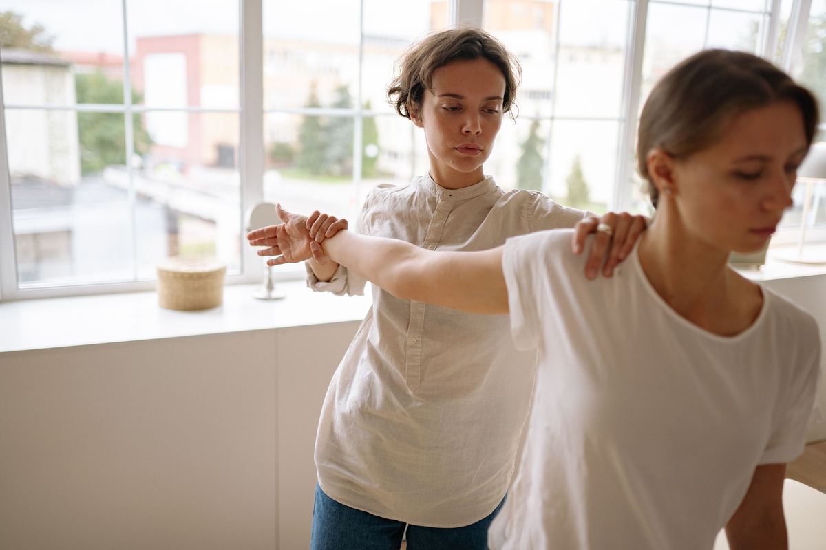 Een kinesitherapeute begeleidt een vrouw bij een oefening voor arm en schouder, bij een raam